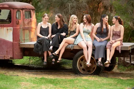 Five young women in black and deep green dresses seated on the rust-red flatbed truck at Premier Studio