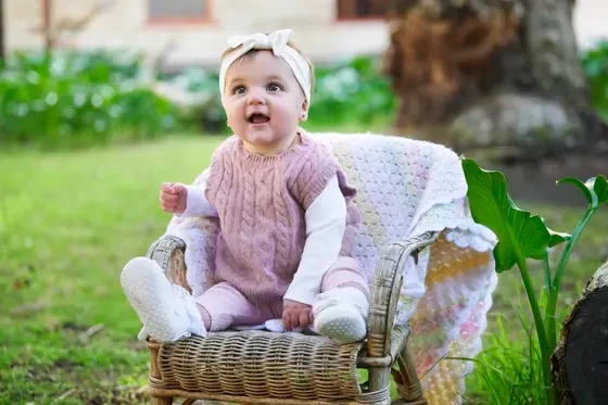 Baby girl in a dusty pink knit vest and cream top sitting in a wicker chair at Premier Studio