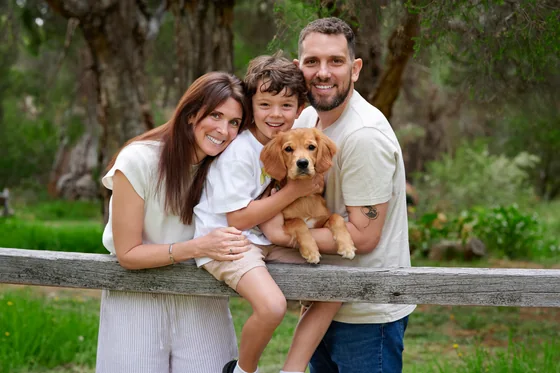 Parents in cream and white with their son and golden puppy at the timber fence at Premier Studio