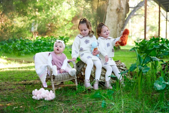 Two little girls in cream and dusty pink knits on a wicker chair with their toys at Premier Studio