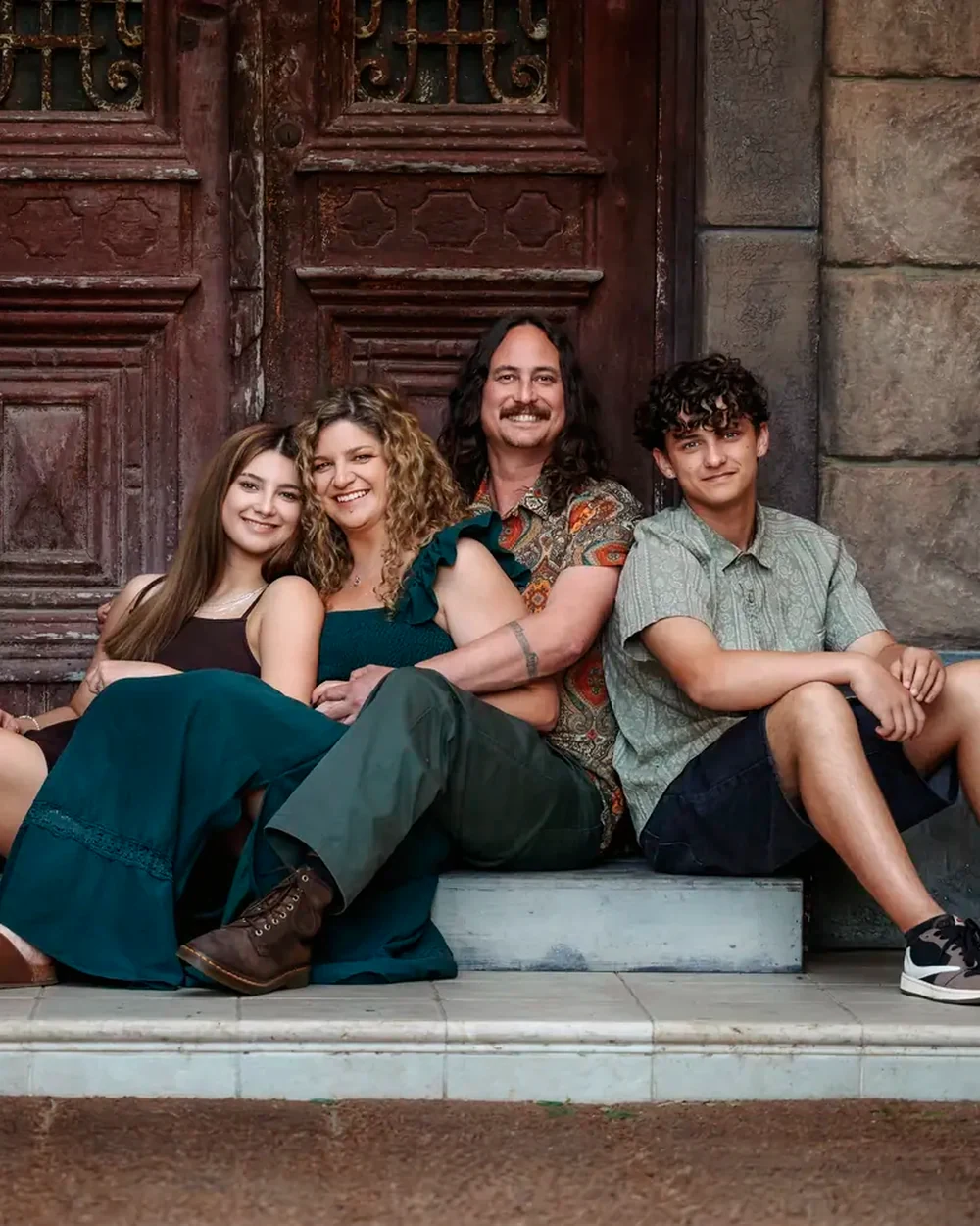 Family of four with mum in an emerald dress seated before the red timber doors at Premier Studio